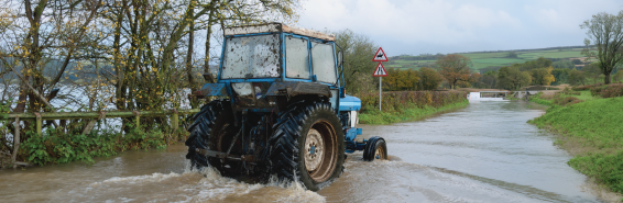Blue tractor driving through floodwater on a rural road surrounded by trees and countryside.